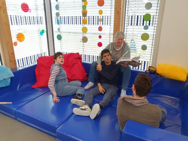 Drei Menschen sitzen auf einem ausgeklappten blauen Sofa auf dem Boden. Eine Frau sitzt auf der Lehne mit einem Buch in der Hand und liest vor.