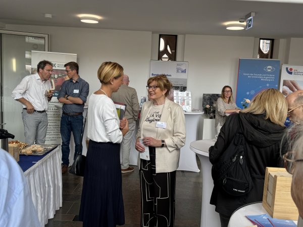 Mehrer Menschen unterhalten sich in einem Foyer. Im Hintergrund befinden sich Stehtische und Roll-ups.