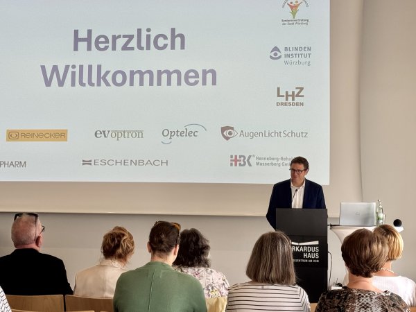 Ein Mann steht vor einem Pult und spricht zu einem Publikum. Hinter ihm ist eine Präsentation projiziert. Die Präsentationsfolie enthält den Text Herzlich Willkommen und mehrere Logos.