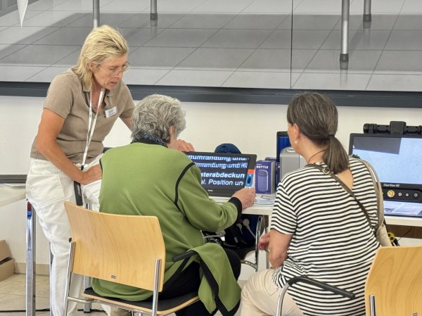 Zwei Frauen sitzen an einem Tisch. Eine der Frauen hat ein Bildschirmlesegerät vor sich und wird von einer weiteren Frau, links von ihr, angeleitet.