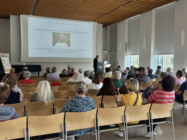 Ein Mann hält einen Vortrag in einem großen bestuhlten Saal. Neben ihm wird eine Präsention projiziert.