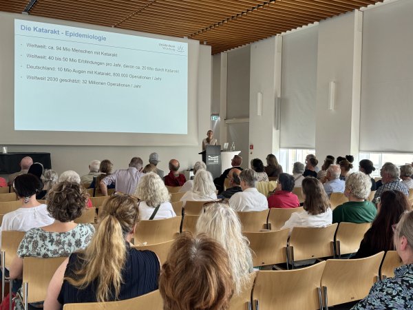 Eine Frau hält einen Vortrag in einem großen bestuhlten Saal. Neben ihr wird eine Präsention projiziert. Die Präsentationfolie enthält die Überschrift: Katarakt - Epidemiologie.