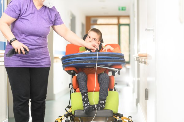 Ein junger Junge sitzt in einem Rollstuhl, der von einer Betreuerin gelenkt wird.
