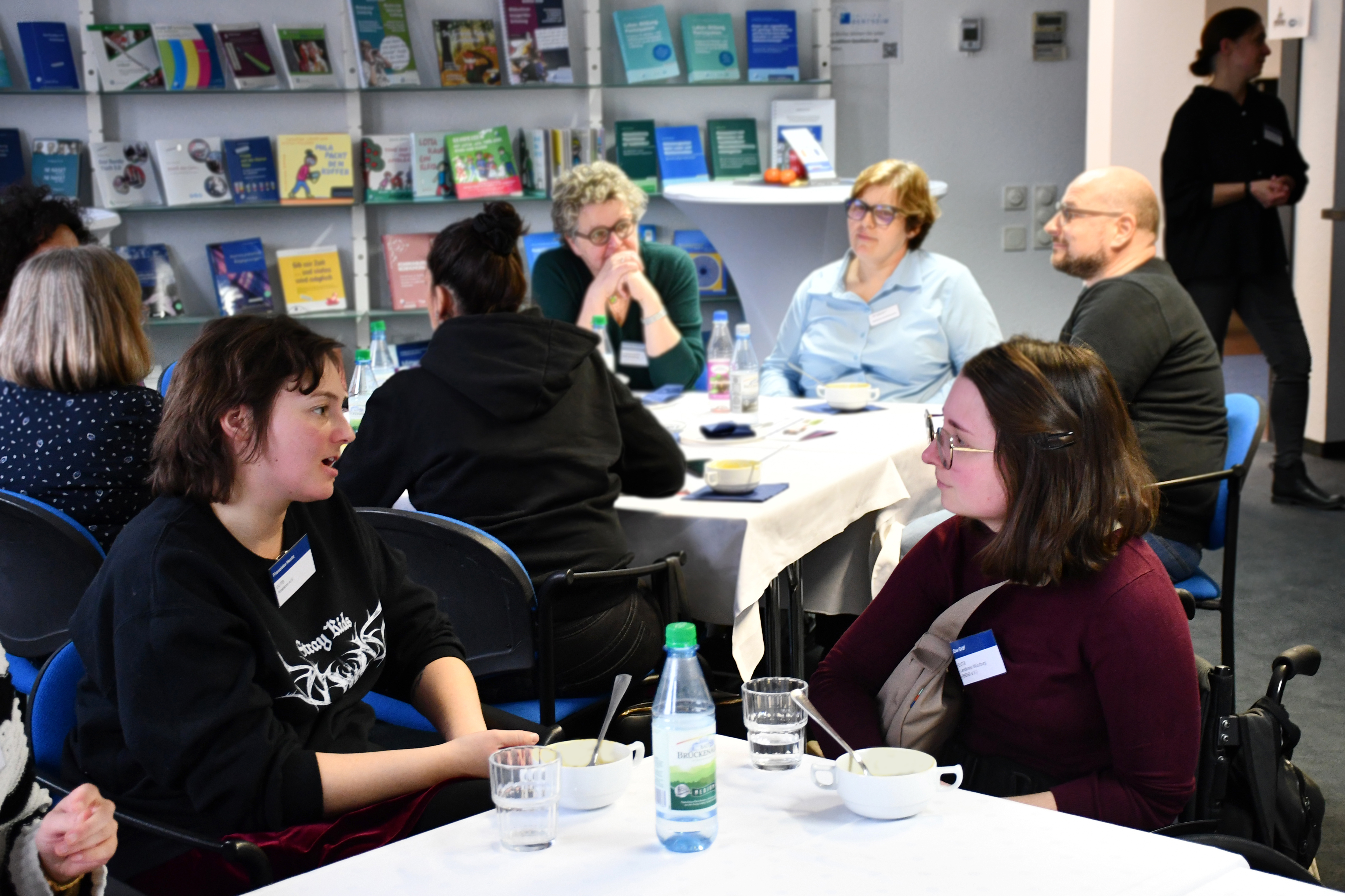 Im Vordergrund unterhalten sich zwei Frauen und sitzen an einem Tisch. Vor ihnen befindet sich eine Wasserflasche und Suppenteller. Im Hintergrund sind mehrere Menschen zu sehen, die ebenfalls an einem Tisch sitzen und sich unterhalten. 