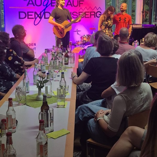 Eine Person spielt auf einer Bühne Akustikgitarre, daneben stehen zwei weitere Personen. Vor der Bühne sitzen zahlreiche Gäste an langen Tischen mit Getränken. Die Bühne ist farbig beleuchtet, im Hintergrund sind große Fenster und ein Banner mit der Aufschrift „Augen auf den Dachsberg“ zu sehen.