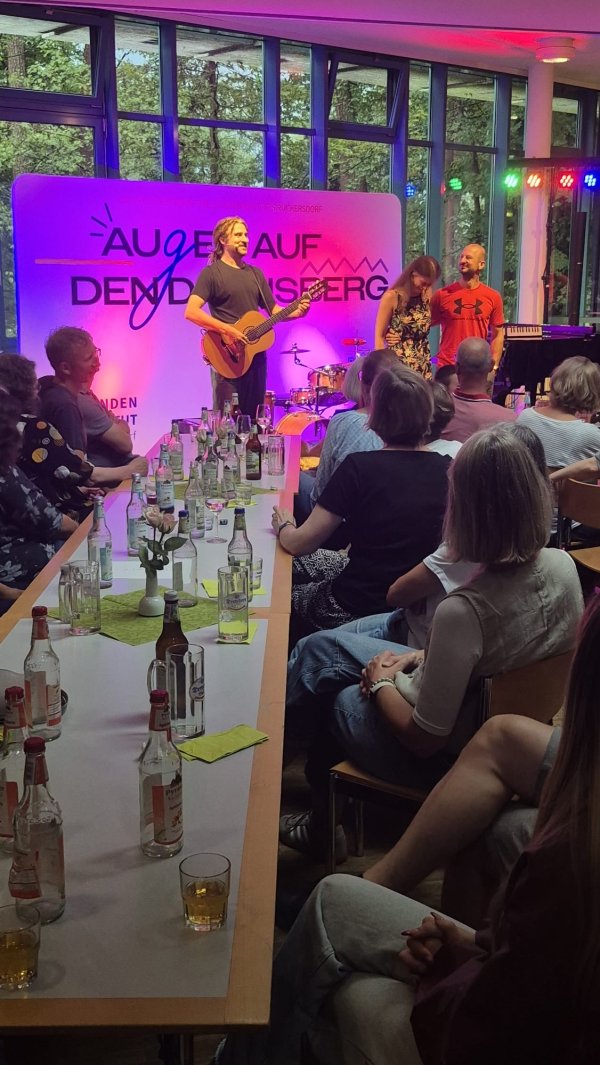 Eine Person spielt auf einer Bühne Akustikgitarre, daneben stehen zwei weitere Personen. Vor der Bühne sitzen zahlreiche Gäste an langen Tischen mit Getränken. Die Bühne ist farbig beleuchtet, im Hintergrund sind große Fenster und ein Banner mit der Aufschrift „Augen auf den Dachsberg“ zu sehen.