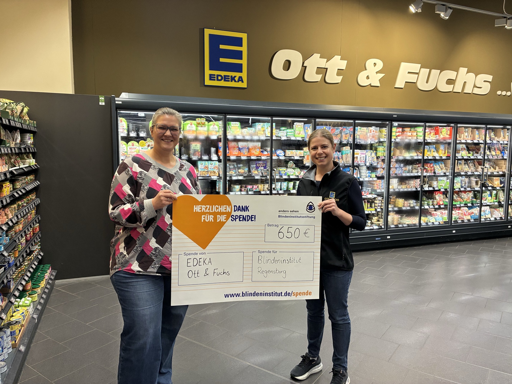 Frau Fuchs und Frau Büchau halten den symbolischen Spendenscheck im Supermarkt Edeka Ott & Fuchs