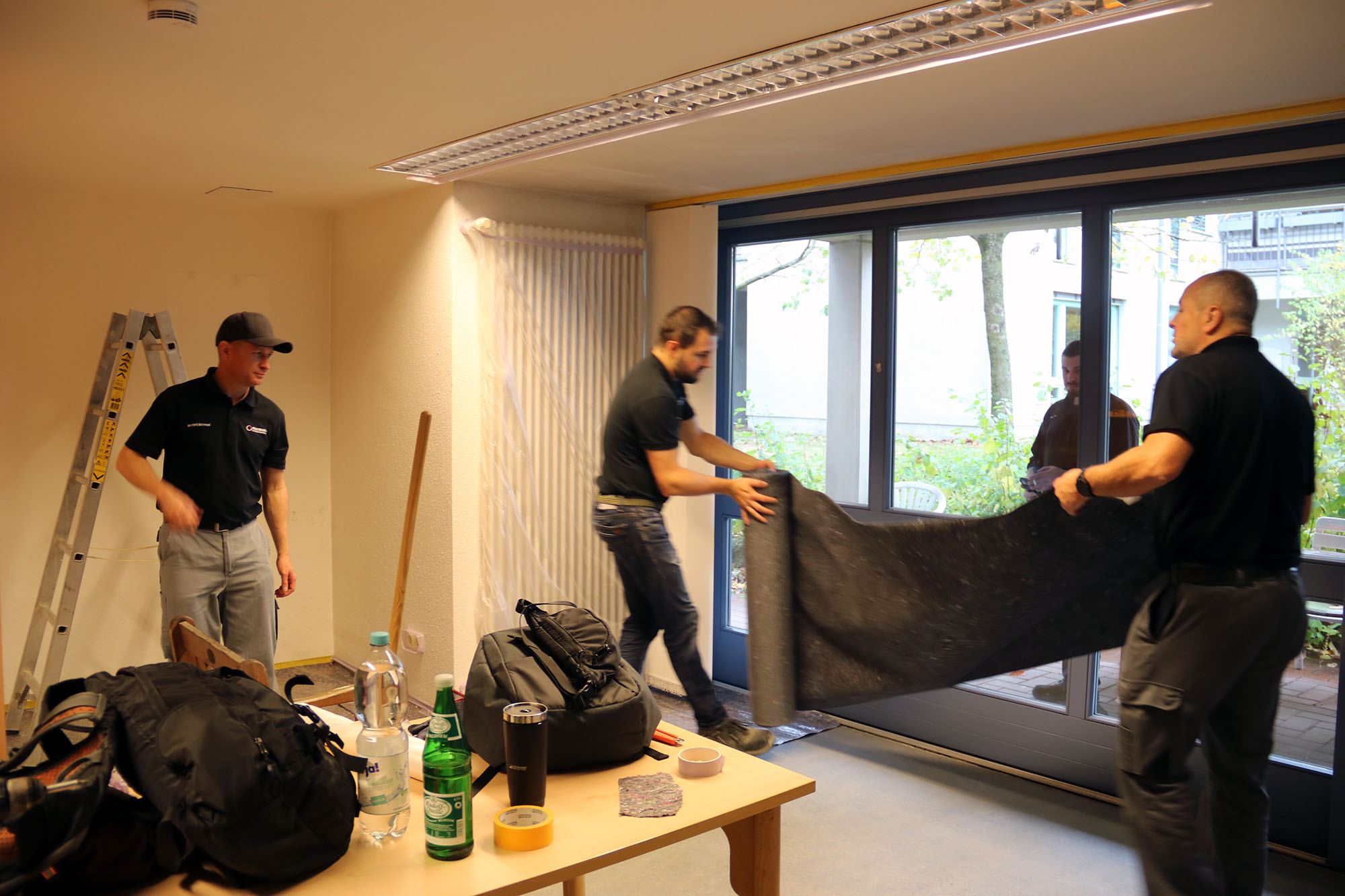 Zwei Männer stehen vor einer Fensterfront und legen eine Malerplane aus. Ein Mann steht links daneben und schaut zu.