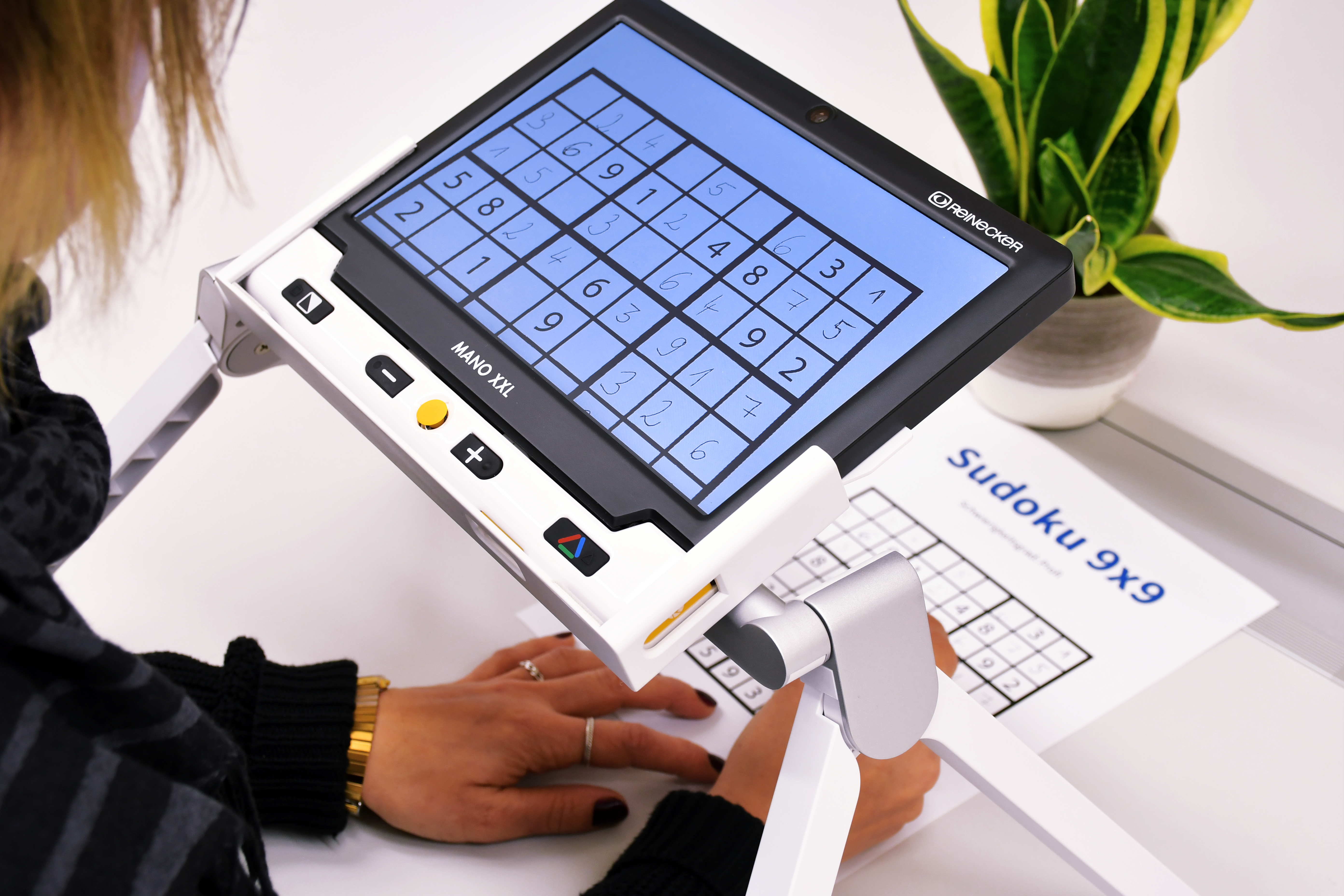 Eine Frau löst ein ausgedrucktes 9x9 Sudoku-Rätsel und nutzt zur Vergrößerung ein Bildschirmlesegerät. Dieses befindet sich auf einem Schreibtisch. 