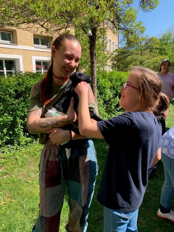 Auf dem Bild sieht man eine junge Frau, die ein Lämmchen hält und ein Mädchen, dass das Lämmchen streichelt,.