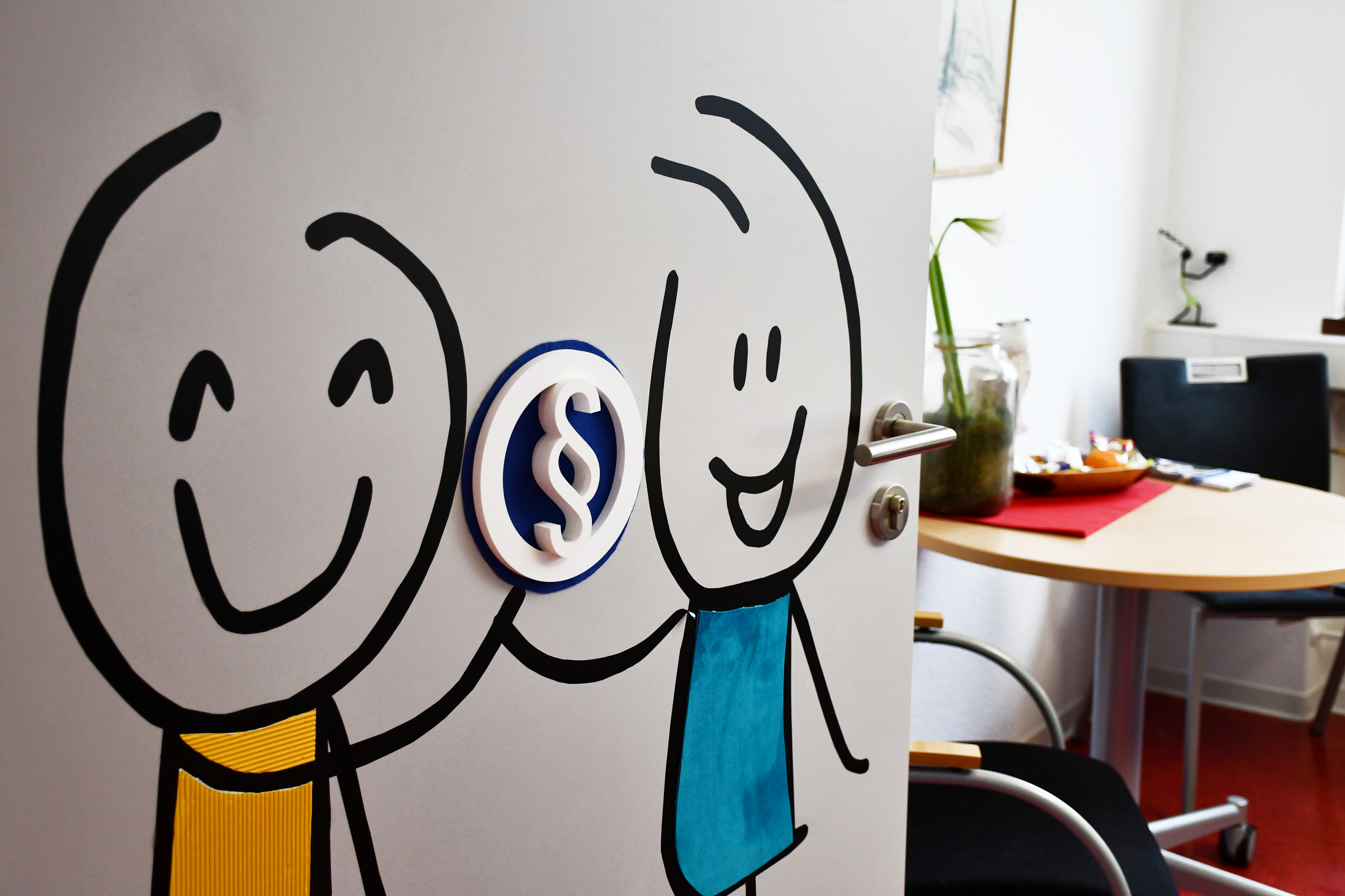 An einer weißen Tür sind zwei bunte Strichmännchen aufgemalt. Sie halten einen Ball in den Händen. Auf diesem ist ein taktiles Paragraphen-Symbol befestigt. Die Tür ist geöffnet und man sieht einen Tisch mit zwei Stühlen. Der Tisch ist weihnachtlich dekoriert. 