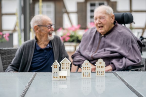 Zwei ältere Männer sitzen an einem Tisch. Einer von ihnen ist im Rollstuhl und trägt ein Hörgerät, der andere eine Brille. Sie schauen sich an und lächeln. Vor ihnen befinden sich kleine Holzmodelle von Häusern.