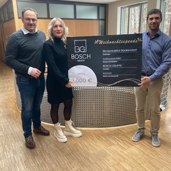 Zwei Männer und eine Frau stehen in einem Raum. Zwei von ihnen halten einen Spendenscheck mit der Aufschrift BG Bosch Gruppe - Weihnachtsspende - Betrag 2000€ - Blindeninstitut Rückersdorf hoch.