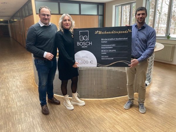 Zwei Männer und eine Frau stehen in einem Raum. Zwei von ihnen halten einen Spendenscheck mit der Aufschrift BG Bosch Gruppe - Weihnachtsspende - Betrag 2000€ - Blindeninstitut Rückersdorf hoch.