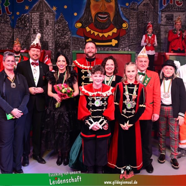 Ein Gruppenbild auf dem Männer, Frauen und Kinder zu sehen sind. Die meisten von ihnen tragen traditionelle Faschingskostüme. Sie alle tragen eine Medaille um den Hals. Eine Frau hält einen Blumenstrauß in den Händen. Unten ist die Aufschrift Fasching ist unsere Leidenschaft - www.gildegiemaul.de zu sehen.