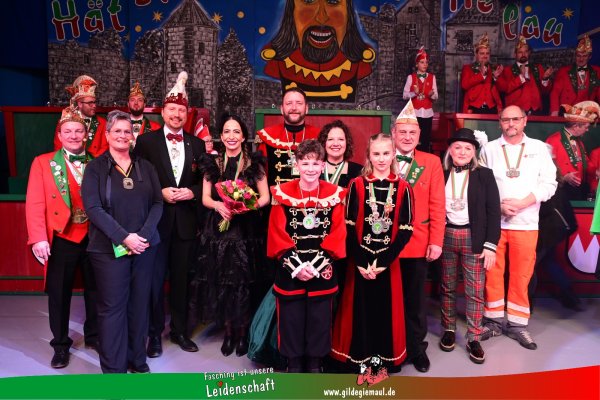 Ein Gruppenbild auf dem Männer, Frauen und Kinder zu sehen sind. Die meisten von ihnen tragen traditionelle Faschingskostüme. Sie alle tragen eine Medaille um den Hals. Eine Frau hält einen Blumenstrauß in den Händen. Unten ist die Aufschrift Fasching ist unsere Leidenschaft - www.gildegiemaul.de zu sehen.