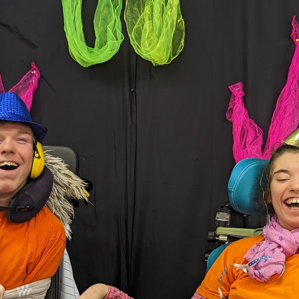 Ein Junge und ein Mädchen lachen in die Kamera. Sie tragen Partyhüte und sitzen im Rollstuhl. Das Mädchen hat Luftschlangen im Haar und der Junge trägt Kopfhörer und ein Nackenkissen. Im Hintergrund ist ein schwarzer Vorhang zu sehen, der mit neonfarbenen Tüchern geschmückt ist.