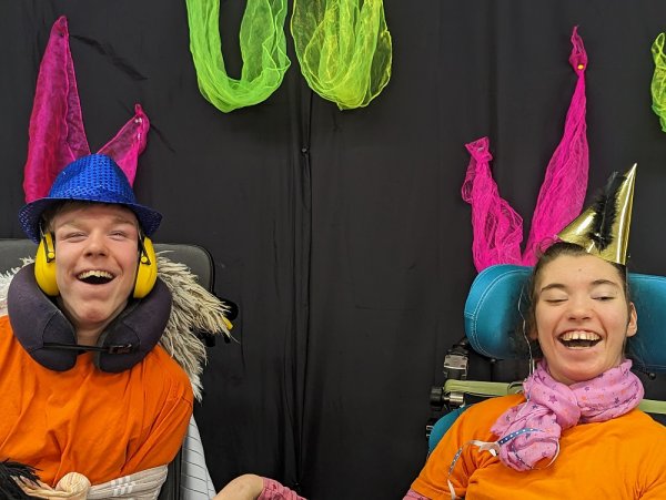 Ein Junge und ein Mädchen lachen in die Kamera. Sie tragen Partyhüte und sitzen im Rollstuhl. Das Mädchen hat Luftschlangen im Haar und der Junge trägt Kopfhörer und ein Nackenkissen. Im Hintergrund ist ein schwarzer Vorhang zu sehen, der mit neonfarbenen Tüchern geschmückt ist.