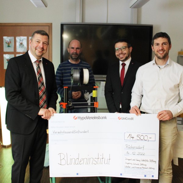 Vier Männer stehen in einem Raum. Zwei von ihnen halten einen großen Spendenscheck mit der Aufschrift Blindeninstitut - 14.500€.