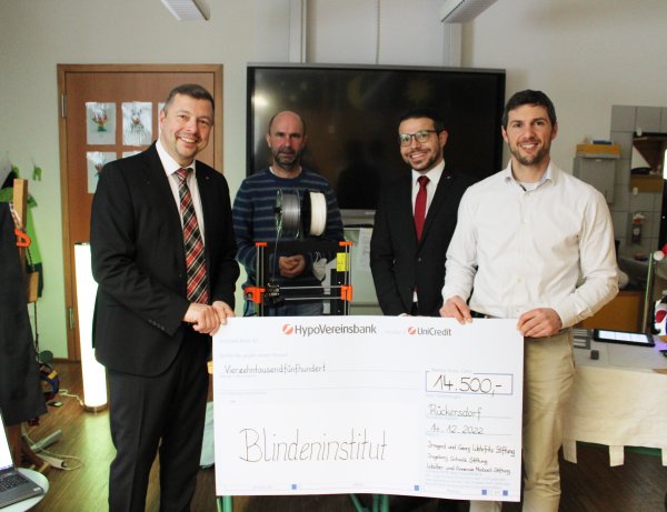 Vier Männer stehen in einem Raum. Zwei von ihnen halten einen großen Spendenscheck mit der Aufschrift Blindeninstitut - 14.500€.