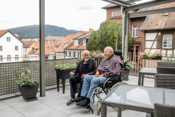 Zwei Männer sind auf einem begrünten Balkon. Eine von ihnen sitzt im Rollstuhl und trägt ein Hörgerät, der andere trägt eine Brille. Beide Männer lächeln in die Kamera.
