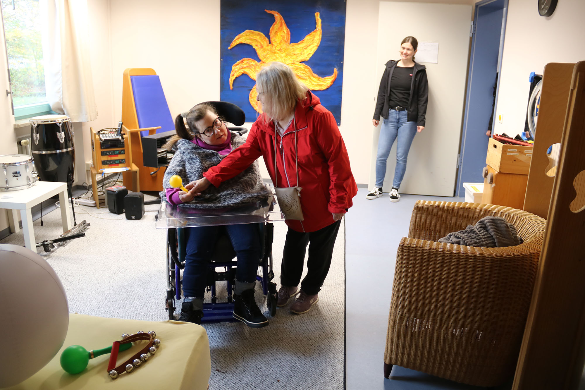 Eine Frau sitzt in einem Rollstuhl und hat eine Rassel in der Hand. Eine andere Frau steht rechts daneben und greift nach der Rassel. Vor den beiden Frauen steht eine Kommode auf der Musikinstrumente liegen