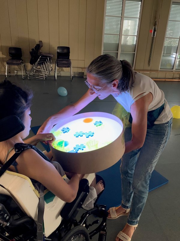 Auf dem Bild sieht man eine Frau und ein Kind im Rollstuhl, die eine beleuchtete BOx mit Gegenstäncden drauf betrachten