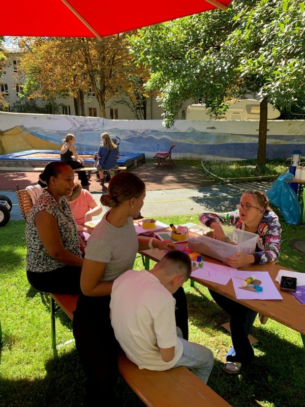 Auf dem Bild sieht man ein paar Menschen, die unter einem Sonnenschirm an einer Biertischgarnitur sitzen.