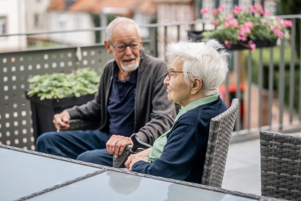 Ein Mann und eine Frau sitzen an einem Tisch auf einem Balkon, der begrünt ist. Sie unterhalten sich.