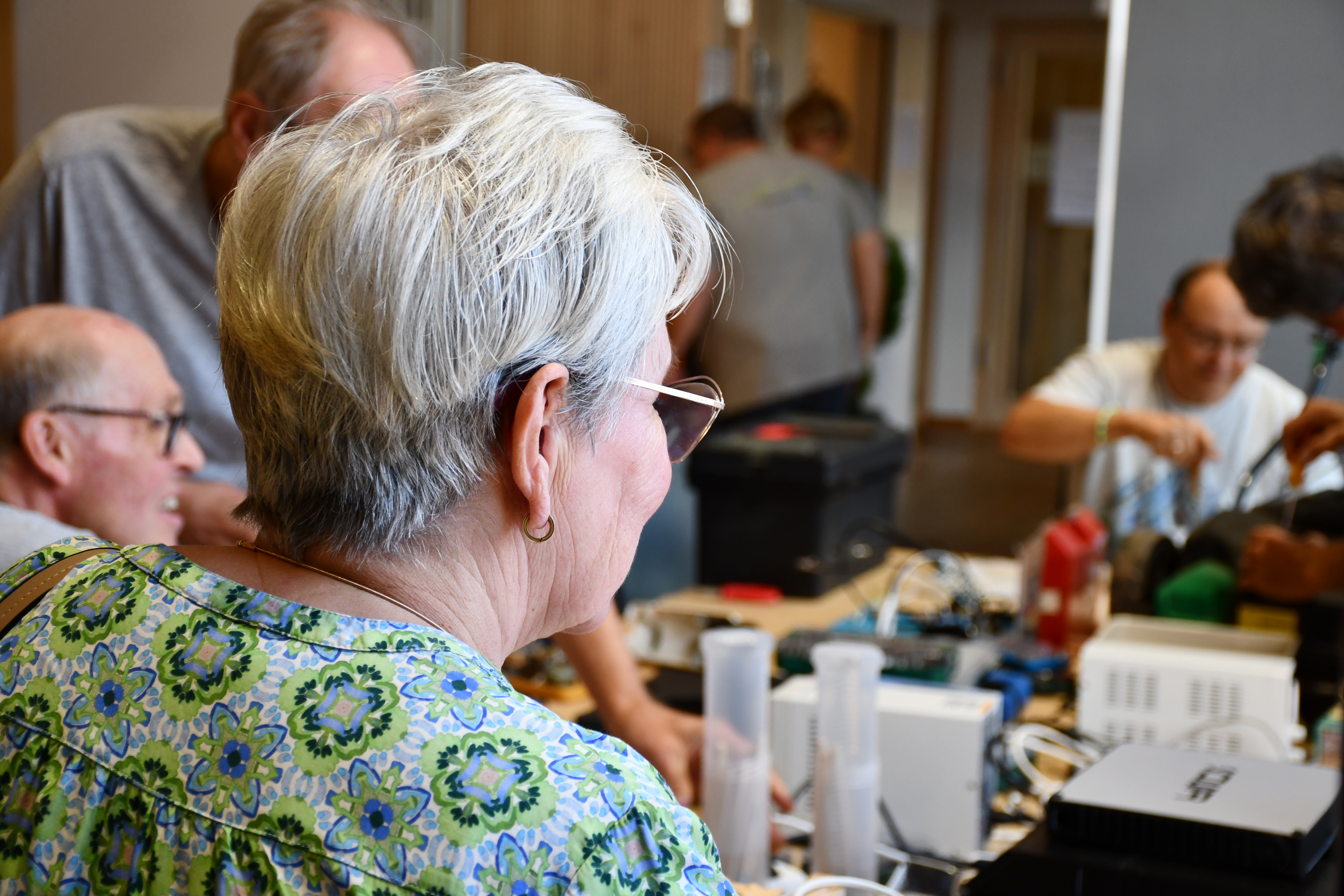 Eine ältere Dame mit Brille schaut auf einen Tisch, an dem mehrere Menschen Gegenstände reparieren. 