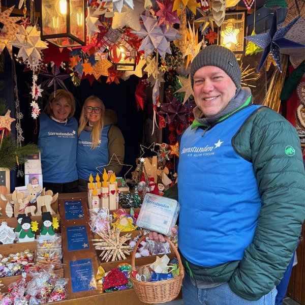 Drei lächelnde Personen stehen an einem weihnachtlich dekorierten Marktstand voller bunter Sterne, Bastelarbeiten und kleiner Geschenkartikel. Sie tragen blaue Westen mit dem Logo der Wohltätigkeitsaktion Sternstunden und wirken einladend und freundlich.
