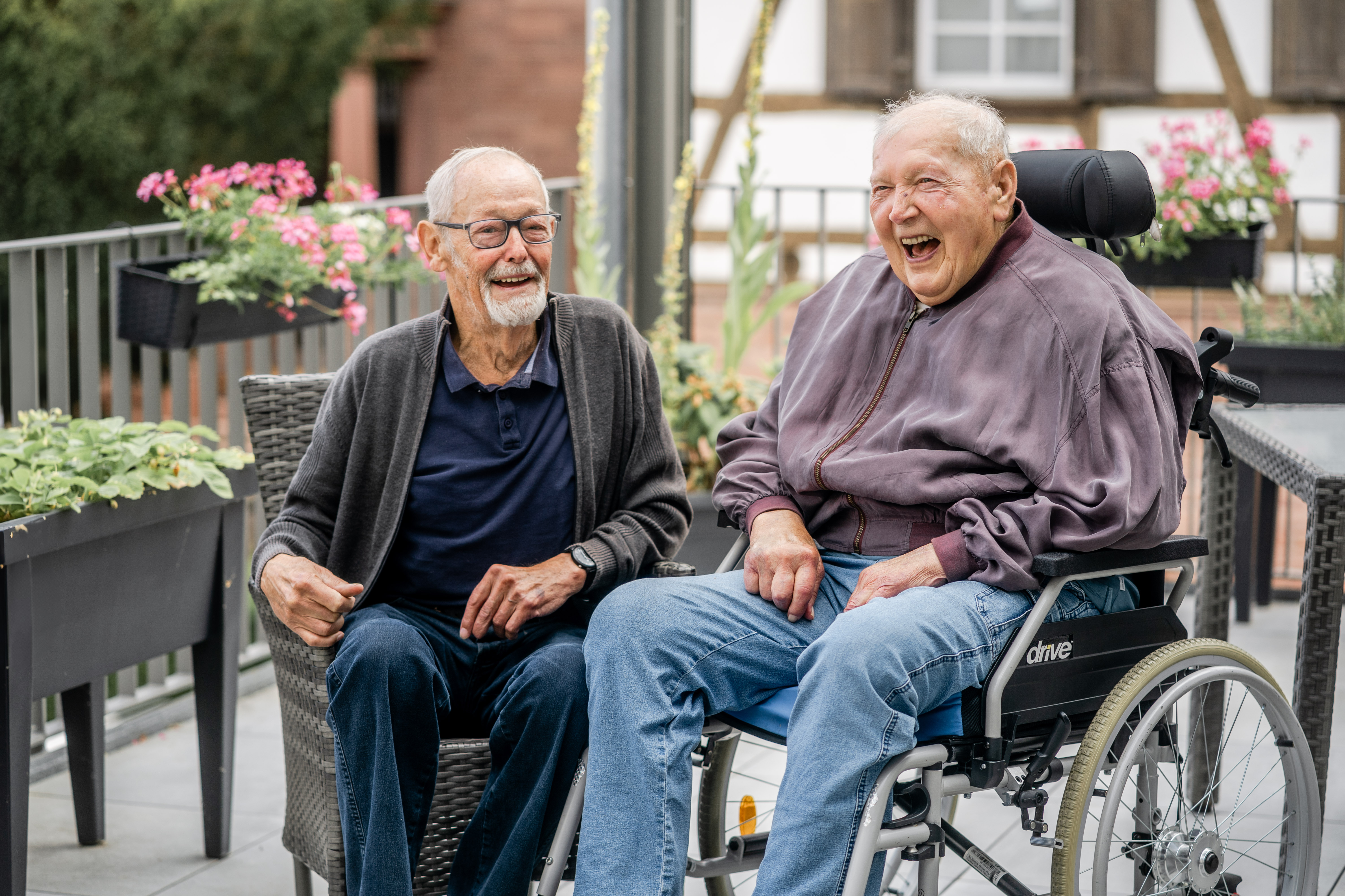 Zwei Männer sind auf einem begrünten Balkon. Eine von ihnen sitzt im Rollstuhl und trägt ein Hörgerät, der andere trägt eine Brille. Beide Männer lächeln in die Kamera. 