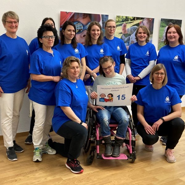Acht Frauen posieren für ein Gruppenbild. Sie alle tragen blaue T-Shirts mit dem Logo des Blindeninstituts. In der Mitte von ihnen sitzt ein Mädchen mit Brille im Rollstuhl. Sie hält ein Bild mit der Aufschrift Herzlichen Glückwunsch zum Jubiläum - 15 in den Händen.