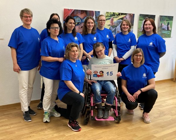 Acht Frauen posieren für ein Gruppenbild. Sie alle tragen blaue T-Shirts mit dem Logo des Blindeninstituts. In der Mitte von ihnen sitzt ein Mädchen mit Brille im Rollstuhl. Sie hält ein Bild mit der Aufschrift Herzlichen Glückwunsch zum Jubiläum - 15 in den Händen.