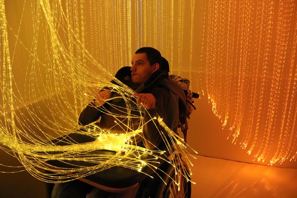 Ein junger Mann im Rollstuhl sitzt vor einem Leuchtvorhang mit kleinen Lichtern