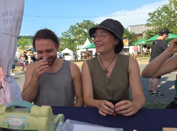 Ein junger Mann und eine junge Frau sitzen an einem Tisch in einem Pavillon und kosten mit geschlossenen Augen Saft