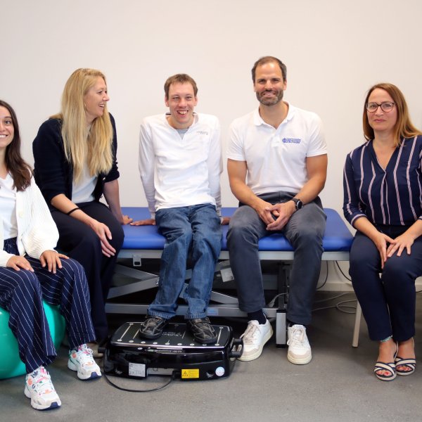 Fünf Menschen sitzen im Halbkreis auf einer Therapieliege und lächeln in die Kamera.