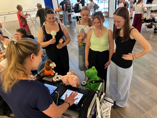 Drei junge Frauen stehen an einem Wagen mit Hilfsmitteln der unterstützten Kommunikation und lassen sich einen digitalen Talker von einer Lehrerin erklären.