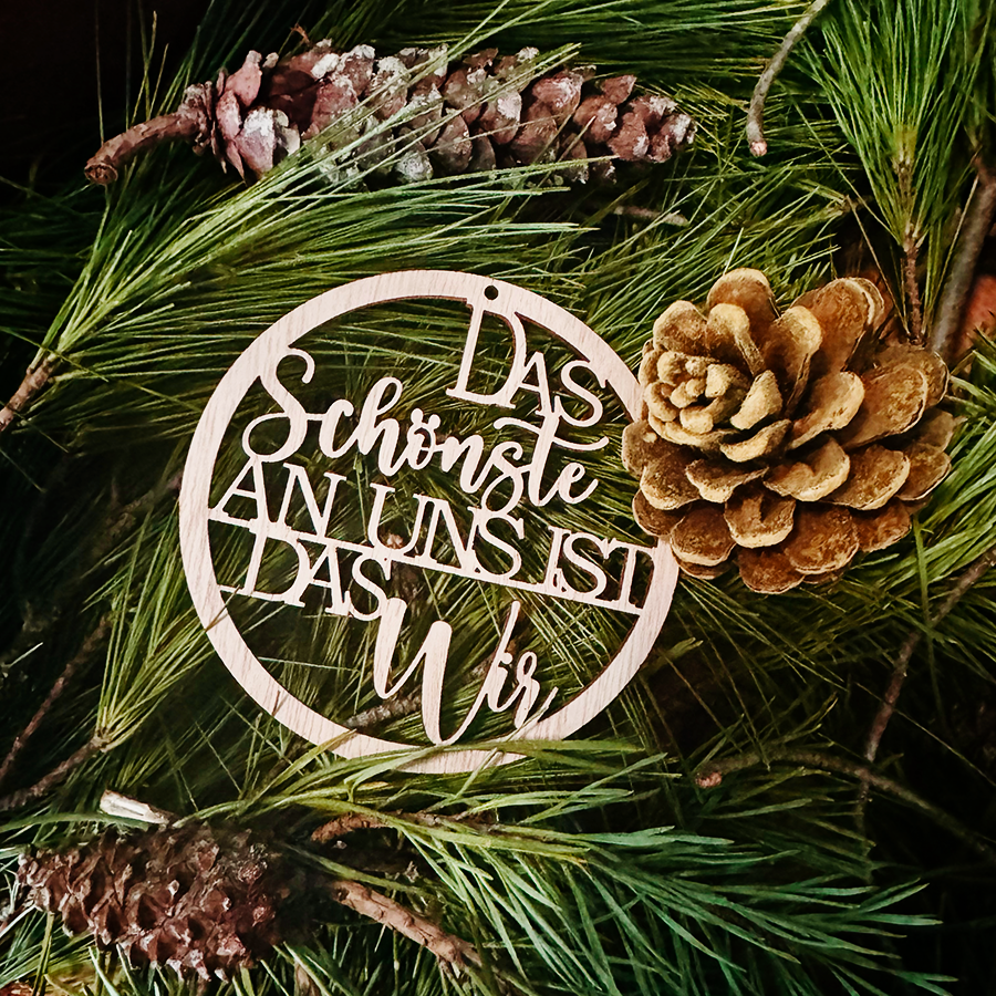 Es ist ein runder Anhänger aus Holz zu sehen, der den Spruch trägt: Das Schönste an uns ist das Wir