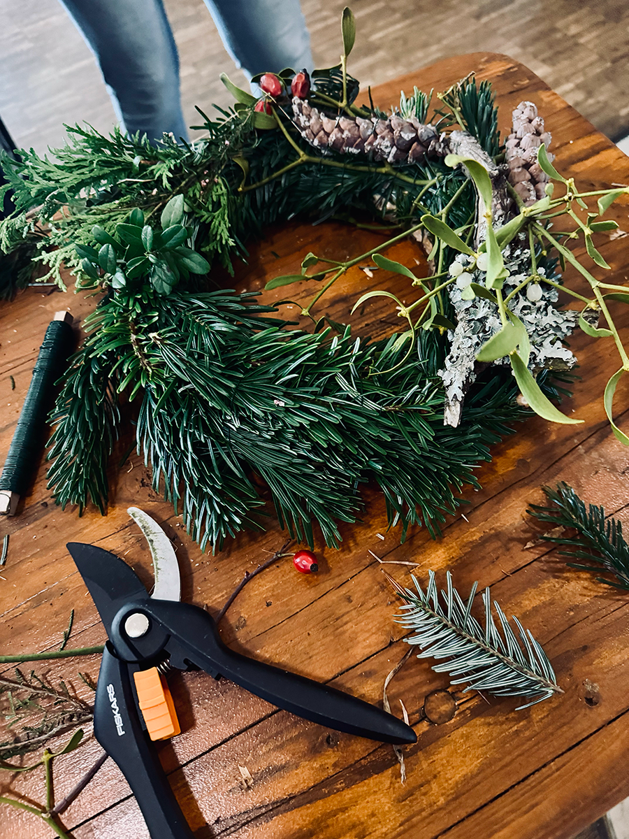 Ein grüner, fertiggestellter Adventskränzen mit saftigen Tannennadeln, roten Beeren, frischen Misteln. Daneben liegen übrige Tannenzweige und eine Gartenschere und Draht.