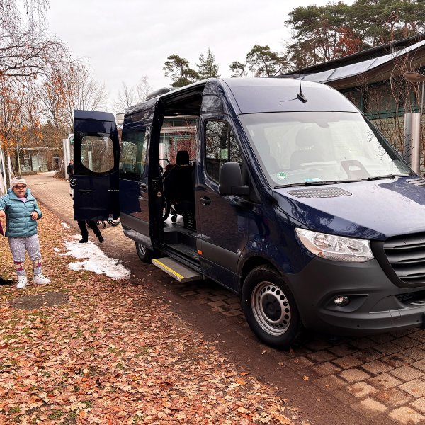 Blauer, barrierefreier Mercedes-Benz Bus mit geöffneter Tür ermöglicht durch die Förderung von Aktion Mensch. Zwei Personen stehen neben dem Fahrzeug, Herbstlaub liegt auf dem Boden.