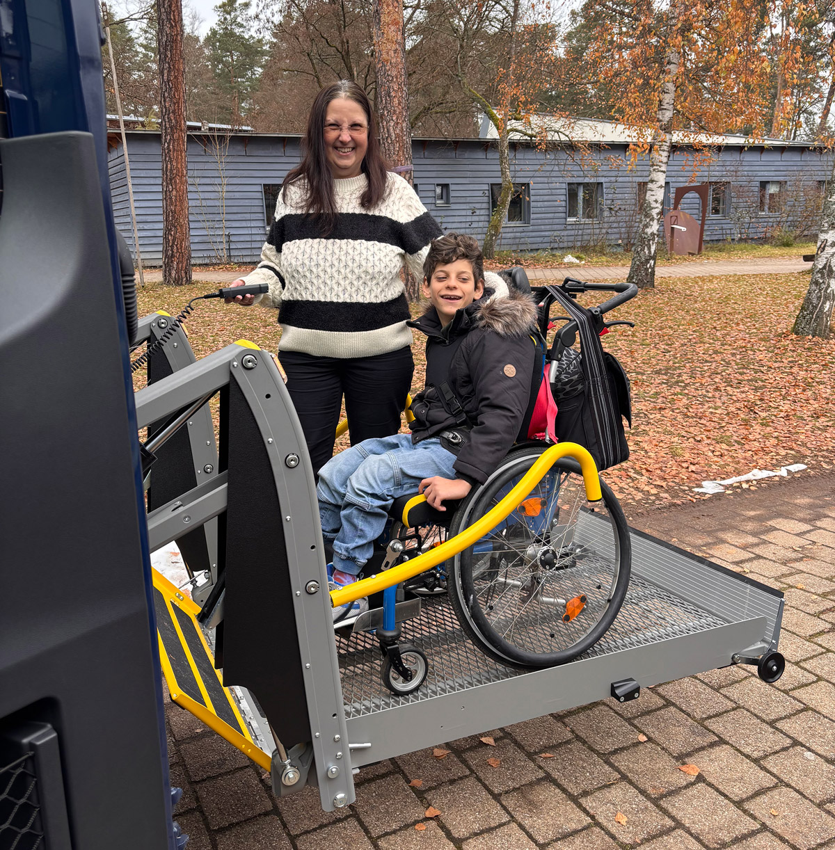 Ein freudiges Kind in einem Rollstuhl steht auf einer ausgefahrenen Hebebühne eines barrierefreien Busses, begleitet von einer weiteren Person. Der Bus wurde durch die Förderung von Aktion Mensch ermöglicht