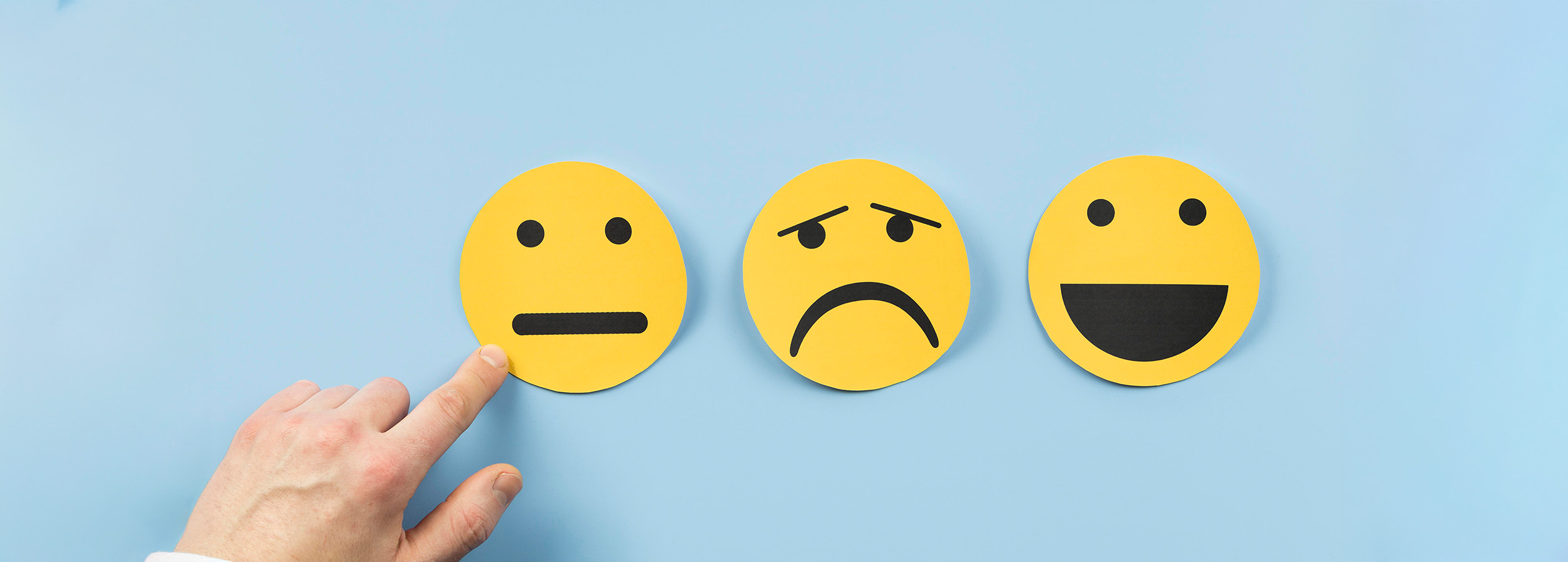 Drei gelbe Smileys auf blauem Hintergrund. Links neutral, in der Mitte traurig, rechts fröhlich. Eine Hand zeigt auf den neutralen Smiley.