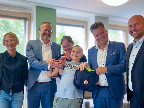 Sechs Personen sind auf dem Bild zu sehen. Ein Junge steht in der Mitte und hält eine kleine, orangene Seepferdchen-Figur in der Hand. Alle lächeln und freuen sich.