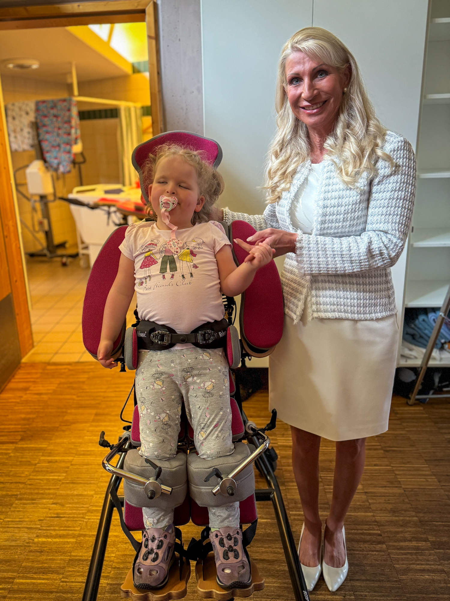 Frau Karin Baumüller-Söder steht neben einem Kind im Stehständer und hält seine Hand.