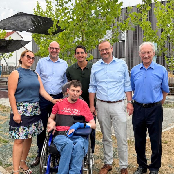 Gruppenfoto im Hof: Sechs Erwachsene stehen hinter einer Person im Rollstuhl; im Hintergrund Bäume, Bänke und ein modernes Gebäude.