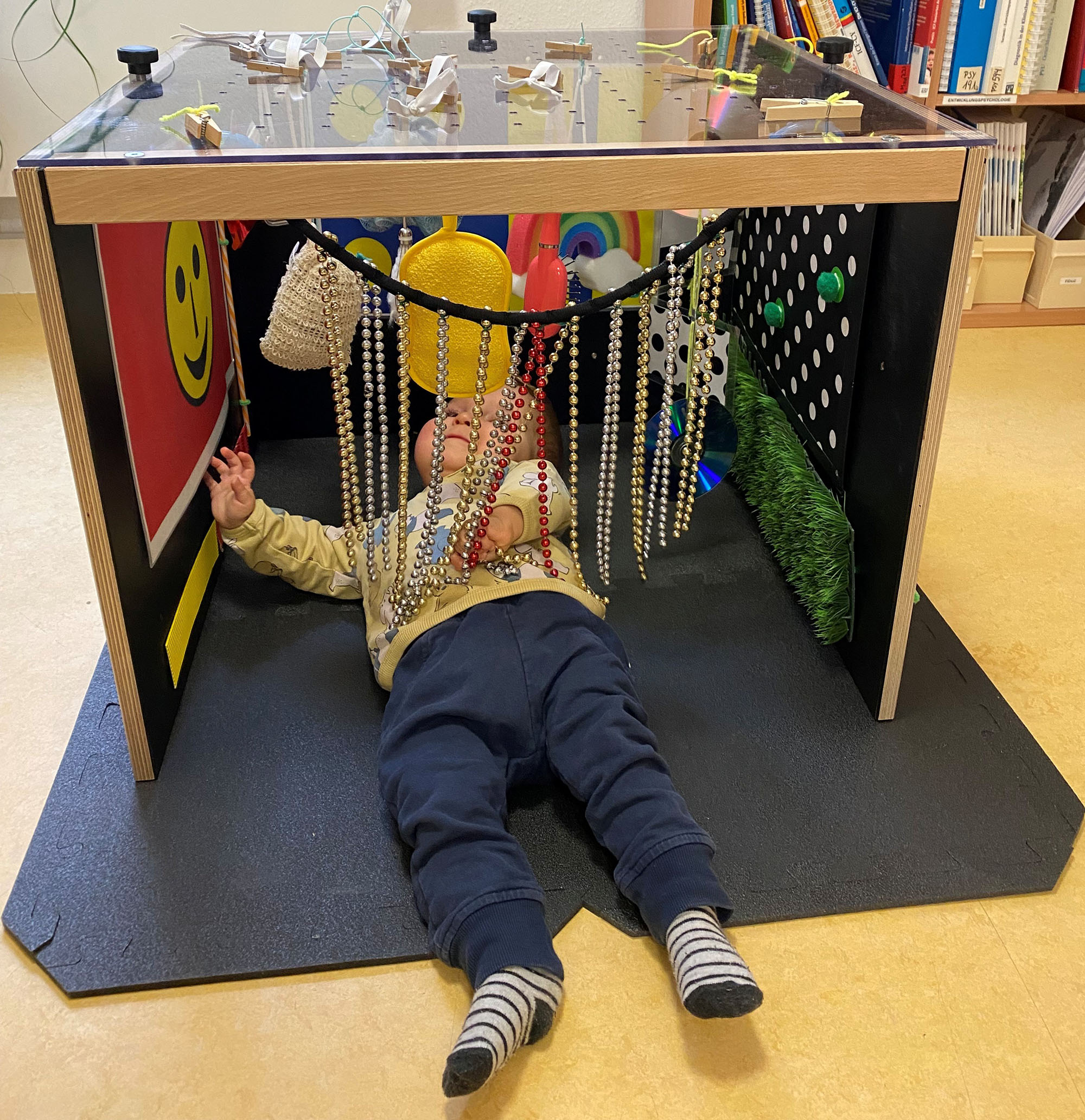 Ein Baby liegt auf einem Teppich unter einer Holzkiste mit durchsichtigem Plexiglasdach. An den Seitenwänden sind bunte Gegenstände befestigt.