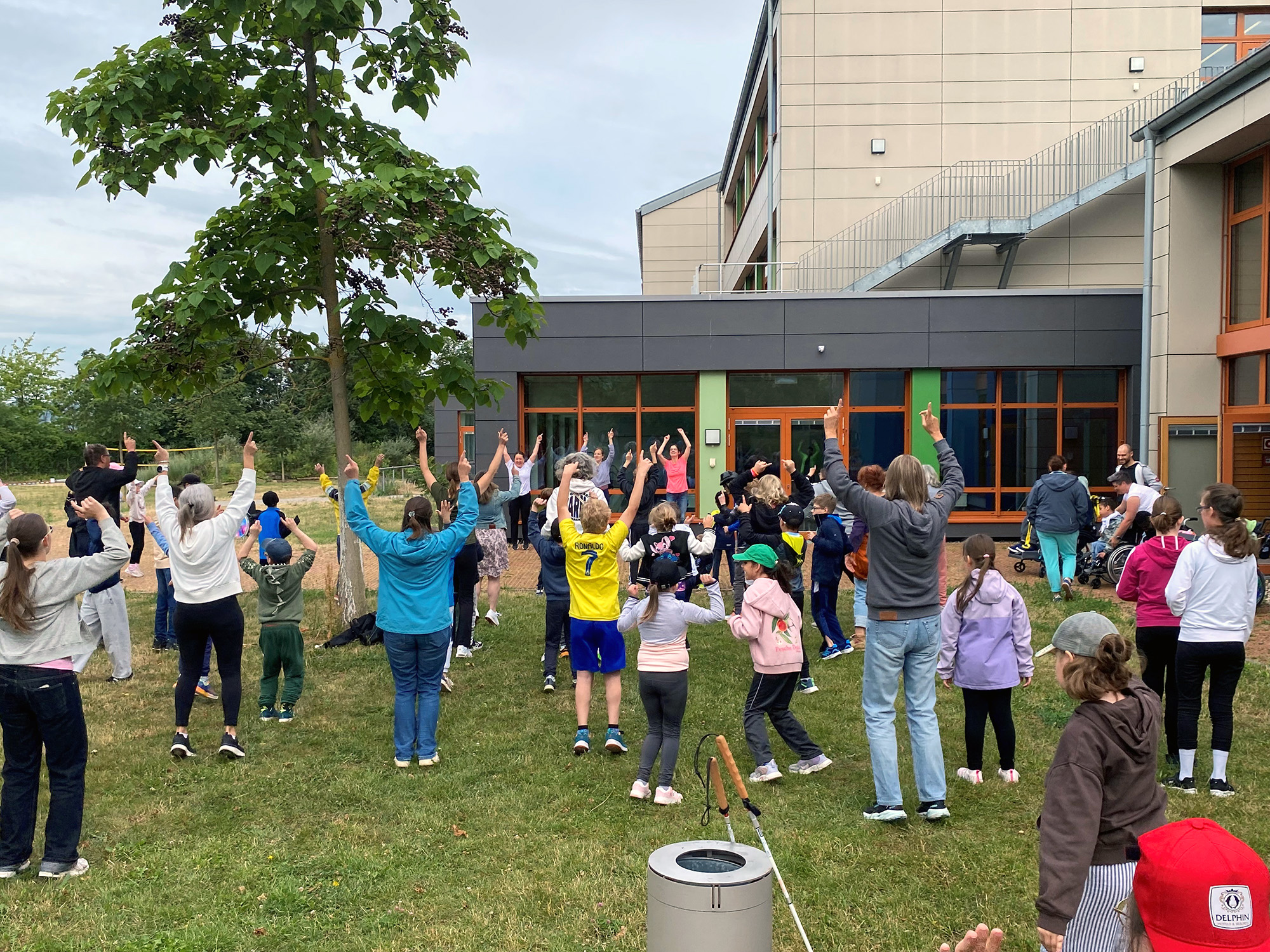Eine große Menge Schülerinnen und Schüler von hinten auf einer Wiese vor einem Schulgebäude - alle strecken die Hände in die Luft. Zwei Frauen sind vor der Menge von vorne zu sehen und strecken die Hände ebenfalls in die Luft..