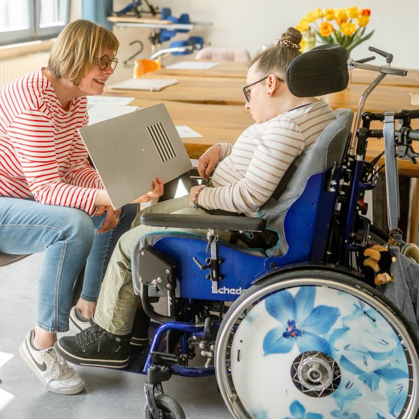 Eine Frau mit Sehtest in der Hand sitzt gegenüber einer Frau im Rollstuhl.