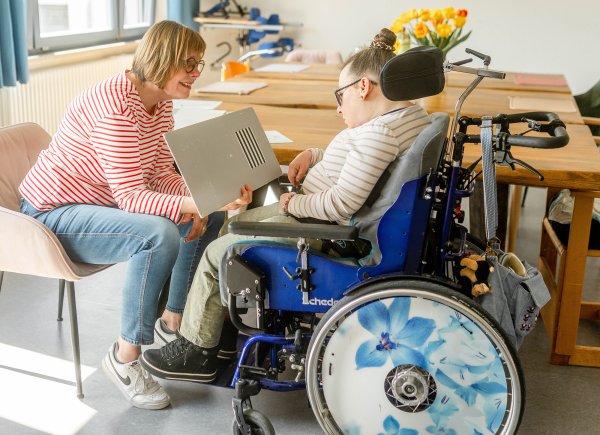 Eine Frau mit Sehtest in der Hand sitzt gegenüber einer Frau im Rollstuhl.